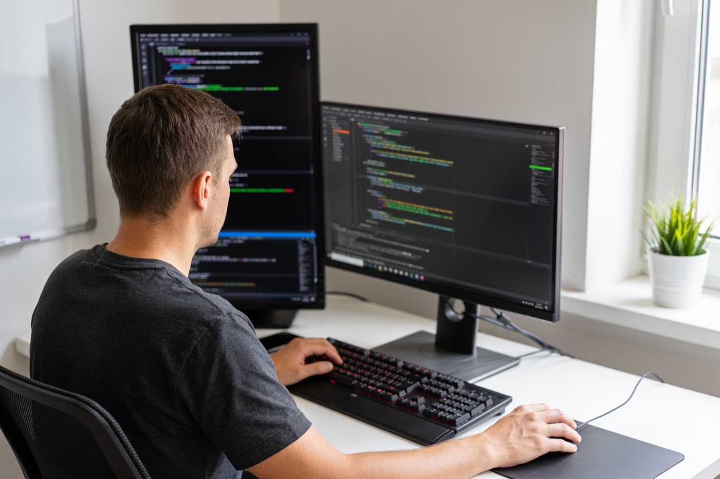 Entwickler von hinten an einem Schreibtisch mit zwei Monitoren, auf beiden läuft ein Code-Editor