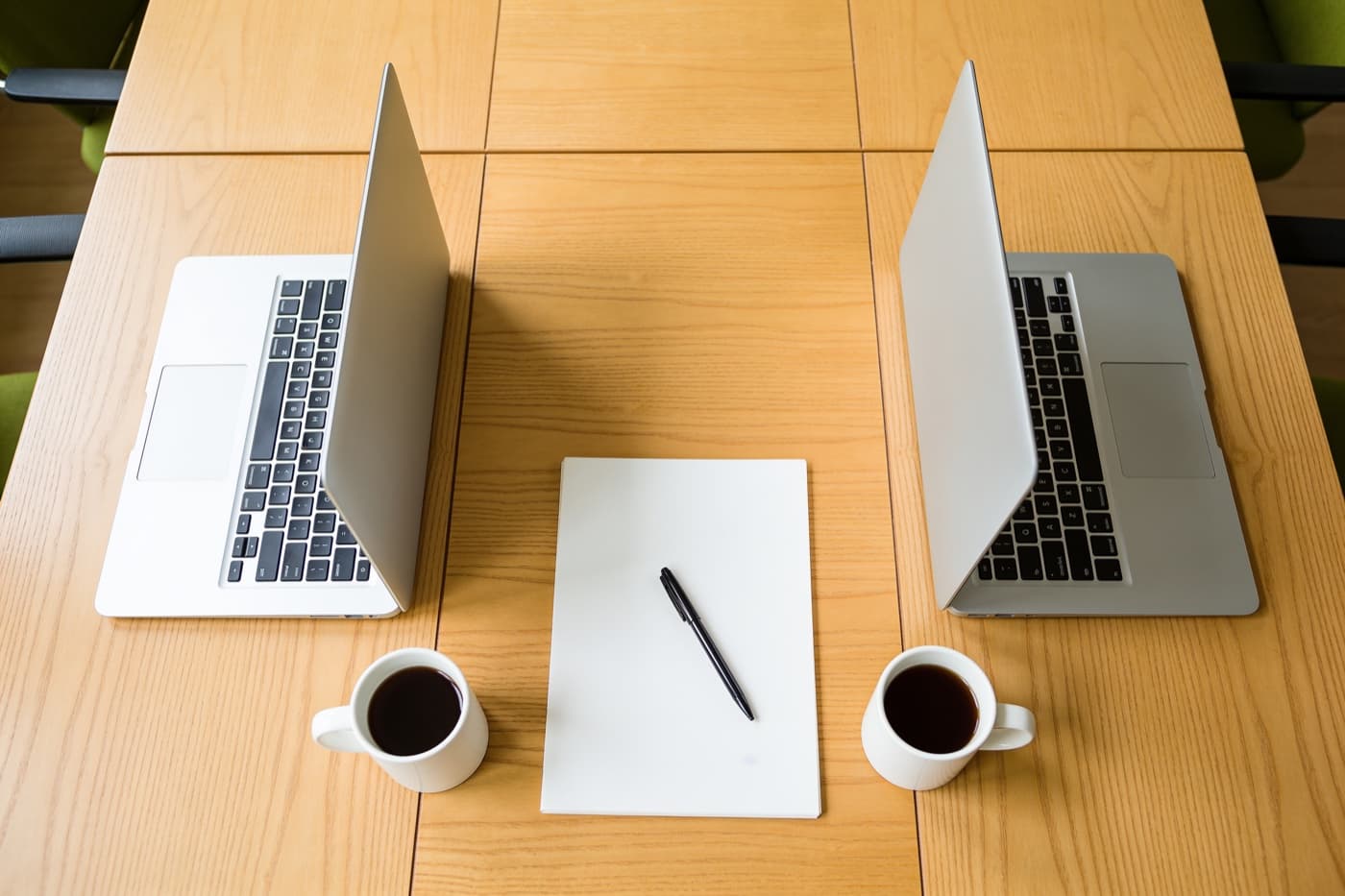 Zwei aufgeklappte Laptops nebeneinander auf einem Holztisch, dazwischen ein leerer Notizblock mit Stift und zwei Kaffeetassen