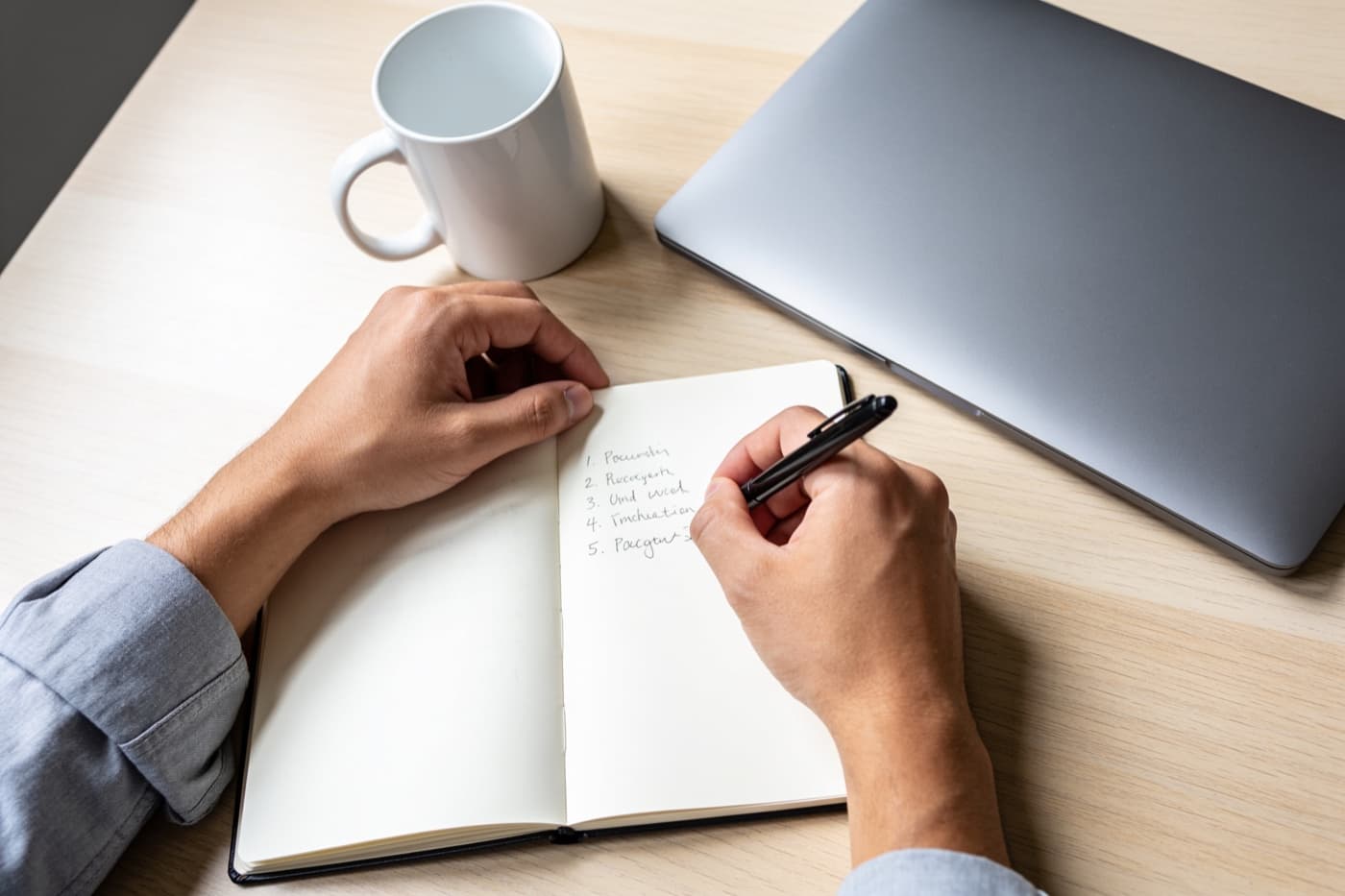 Zwei Hände notieren von Hand eine nummerierte Liste in ein aufgeschlagenes Notizbuch, daneben ein zugeklappter Laptop und eine weiße Kaffeetasse auf einem hellen Schreibtisch