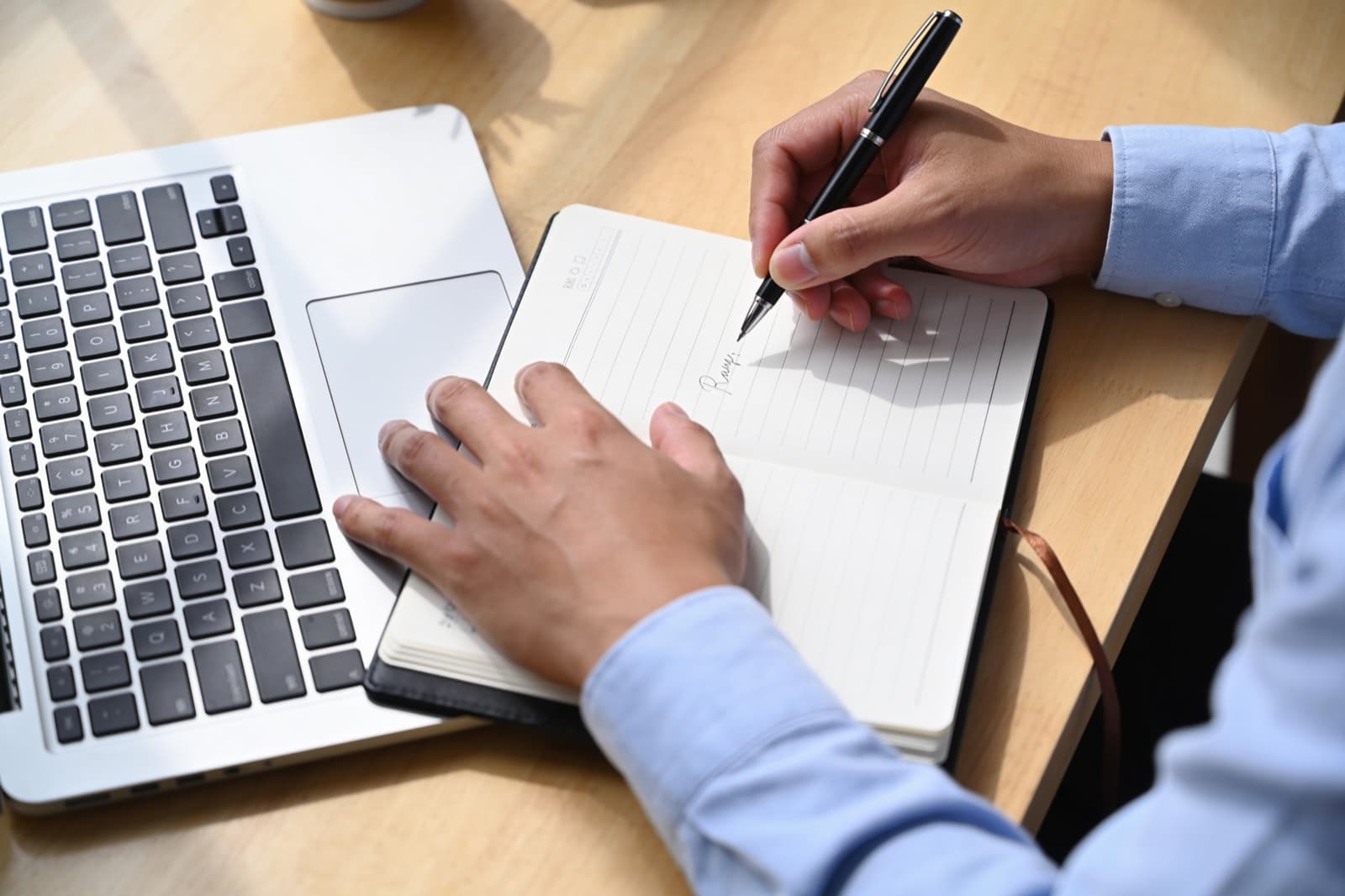 Hände mit Stift über einem Notizbuch neben einer Laptop-Tastatur